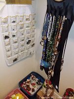 Wide shot including white hanging pocket organizer with earrings, blue hanging jewelry holder with necklaces, and boxes with rings and bracelets below.
