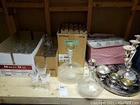 Three boxes of assorted drinking glasses including 21 Waterford Crystal pieces and two glass decanters on wooden shelf.