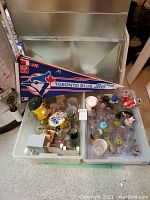Wide shot of all items including Toronto Blue Jays pennant, novelty collectibles, glasses and cups organized in plastic bins and a metal container behind.