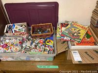 Plastic storage bin with dark red lid removed revealing assorted button pins, small wicker basket of pins, cardboard box of comics and stack of comics.