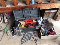 Wide view of plastic toolbox with red interior containing hand tools, assortment of combination wrenches on the floor, and a box of power tools with circular saw blade