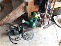 Photo showing the lot of garden items on concrete floor in garage: electric hedge trimmer, watering can, garden rakes, hand tools, fertilizer, and decorative sun face ornament.