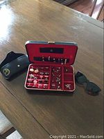 Photo showing red velvet lined case open with over 10 pairs of men's cufflinks arranged inside, along with several tie clips, and a pair of black Ray-Ban sunglasses with case on wooden table.
