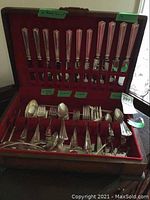 Flatware set displayed in an open red velvet-lined case, showing cutters with distinct elongated geometric handles on upper lid and assorted spoons and forks in the bottom section.