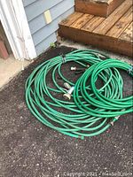 Two coiled green garden hoses with attached nozzles laid on pavement and a concrete/wood step background