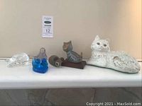 Wide view showing all five figurines on a mantle shelf: large white cat lying down, blue glass cat, clear glass mouse, blue bird, small gray cat on wood base.