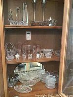 Wood cabinet showcasing various glass and crystal serveware including bowls, candy dishes, salt and pepper shakers, and a glass basket with handle.