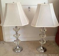 Pair of metal and Lucite base table lamps shown side by side with white fabric shades. Lamps stand on carpeted floor against a white background.