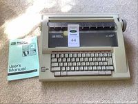 Top view of beige Smith Corona DXL 100 electric typewriter with keyboard and closed platen cover alongside teal user manual.