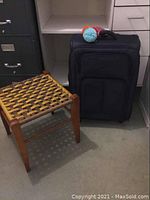 Black Delsey suitcase placed next to a wooden woven stool on carpeted floor