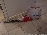 Full view of the red electric hedge trimmer lying on floor, showing cutting blade, handle, power cord and clipping collection bag.