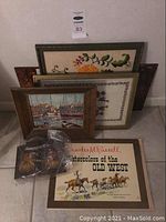 Four framed vintage artworks stacked against a wall including embroidered floral pieces, a harbor painting, and a Charles M. Russell print.