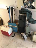 Photo showing folding golf cart with attached green golf bag, red and white Playmate cooler, carriers, and golf balls on floor.