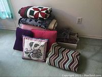 Assorted bedding and pillows stacked on the floor including quilts, blankets, and pillows of various sizes and colors.