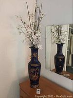 Full view of tall blue decorative vase with artificial stems beside a mirror.