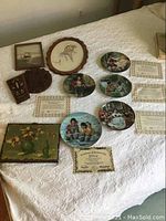 Collector plates and certificates arranged on bed showing multiple artistic scenes and accompanying paperwork