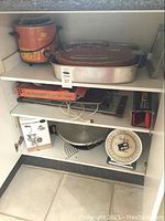Shelf with Sunbeam Heritage Pot crock pot, electric roaster, manual meat grinder box, kitchen scale, mandoline slicer, and french fry cutter inside cabinet