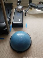 Blue half aerobic balance ball with black base, positioned on carpeted floor near workout bench.