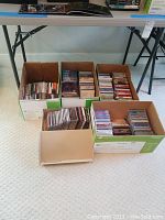 Five cardboard boxes containing a large number of assorted music CDs in jewel cases, placed on floor below table.