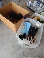Photo showing cardboard box with several pairs of boots and cleats inside and a clear plastic container with more boots and footwear including blue overshoes and fleece-lined boots.
