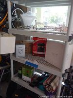 Photo showing white plastic shelving unit with multiple items including Honeywell heater box, other car maintenance product boxes, and assorted items on lower shelf. Part of bicycle wheel visible in background.