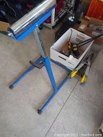 Photo of blue Kobalt roller stand with adjustable height next to box containing C-clamps and yellow handle metal shears and other tools on concrete floor.