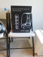 Box packaging for Harman Kardon SoundSticks III speaker system showing product name and image, sitting on a chair.