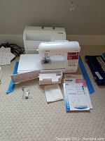 Front view of Brother computerized sewing machine with LCD and keypad, along with included manuals and accessories arranged on floor.