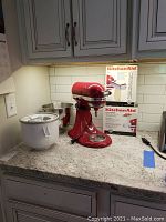 Red KitchenAid mixer on countertop with stainless bowl and two boxed attachments behind