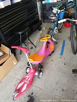 Blue and white Cannondale bicycle with training wheels and basket, pink and orange toddler scooter, red Radio Flyer three-wheel scooter arranged together in garage