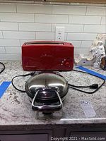 Red KitchenAid toaster with control knob and stainless steel All-Clad waffle maker on kitchen counter.