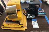 Wide photo showing Nikon lens box, Kodak camera accessories, Polaroid camera and Magicubes flash bulbs box on a table