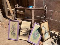 Vintage wood shelf with decorative carved sides against a brick wall with four framed paintings leaning against it outdoors.