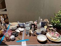 Overview of collection of vintage porcelain and glass items including cups, plates, vases, and decorative pieces arranged on table outdoors.