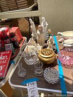 Full view of table showing porcelain figurines, glass decor including crystal bowl, pink bowl, gold decorative item, and Coca-Cola collectibles.
