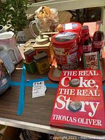 General overview of the lot showing Coca-Cola collectible tray, bottles, red tin bucket, glass pitcher, and the book titled 'The Real Coke The Real Story'