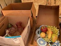 Boxes containing assorted faux fruit, clear glassware, decorative Wedgwood plates and creamer, and other decorative items
