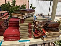 Table filled with a large collection of vintage and antique books mostly stacked in several piles. Visible books include red softcovers and beige hardcover sets.