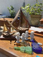 Wide view showing two metal sail boats, four composer bust figurines, blue glass guitar, and glass bottles on table outdoors.