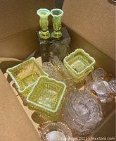 Box containing vintage glassware assortment including yellow-green colored glass candle holders, square dishes, and clear glass items with intricate patterns.