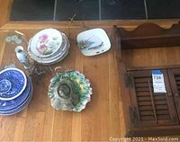 Overview of the lot with all plates stacked in sets along with the Majolica dish, candle holder, and wooden decorative piece on a wooden floor.