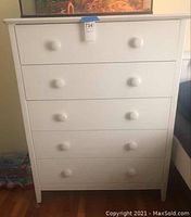 Front view of white dresser showing four drawers with two round knobs on each drawer. The dresser is placed on a wooden floor next to a blue sofa and a low table with bottled water package in background.