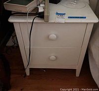 Front view of white two-drawer nightstand showing drawers and knobs with power cords on top.