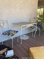 Two white iron chairs with floral cushions positioned at either side of a rectangular white iron table with glass top, outdoor setting on wooden deck, showing some rust and weathering.