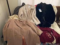 Lot of vintage coats including a light beige faux fur coat, pink double-breasted wool coat, black faux fur coat, and maroon coat with embroidered crest.