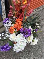 Artificial flower arrangement featuring orange flowers, purple clusters, white flowers, pink buds, tall red flowers, and green spiked foliage.