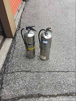 Photo showing two large silver-colored fire extinguishers placed on asphalt surface near a storage unit door.