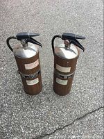 Photo of two large metal fire extinguishers standing side by side on asphalt. Each extinguisher has black trigger handles and brown tanks with labels and gauges.