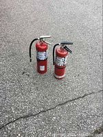 Two medium-sized red fire extinguishers with labels, one with hose attached, placed outdoors on pavement surface.