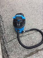 Full view of the blue Dura Vac vacuum cleaner with attached black hose and extended power cord laid on asphalt surface.